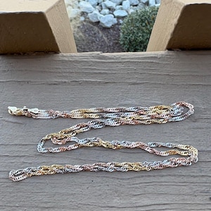 May include: A delicate necklace featuring a tri-color rope chain design. The chain is composed of interwoven strands in gold, silver, and rose gold tones. The necklace is displayed on a weathered wooden surface.