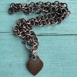 May include: A silver-toned necklace featuring a thick chain made of interlocking circular links and a toggle clasp. A smooth, heart-shaped pendant hangs from the chain, with the number 925 clearly engraved on the small bail at the top of the heart.