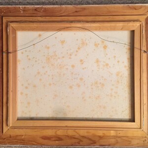 May include: The back of a framed painting with a light brown wooden frame. The canvas is a light beige color with some brown stains.