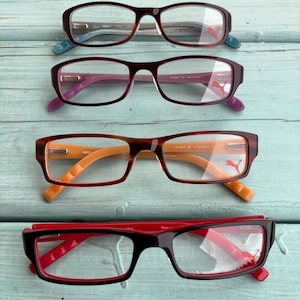 May include: Four pairs of eyeglasses with rectangular frames in various colors: brown, purple, and black. Each pair features a Puma logo on the temple. The frames have different colored accents, including blue, purple, orange, and red.