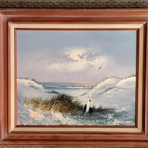 May include: A framed oil painting depicting a seascape with a lone figure in a white hat sitting on a sandy dune. The sky is a pale blue with white clouds and a few birds flying overhead. The ocean is a deep blue with white waves crashing on the shore.