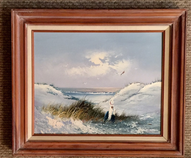 May include: A framed oil painting depicting a seascape with a lone figure in a white hat sitting on a sandy dune. The sky is a pale blue with white clouds and a few birds flying overhead. The ocean is a deep blue with white waves crashing on the shore.