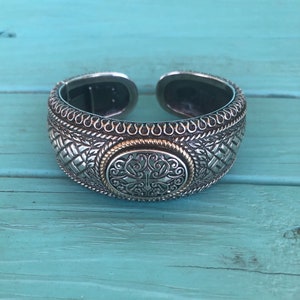 May include: Silver and gold tone cuff bracelet with an intricate design. The bracelet has a woven pattern and a large circular centerpiece with a floral design.
