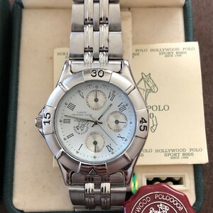 May include: A silver wristwatch with a white face and black Roman numeral hour markers. The watch has three subdials, a date window, and a silver metal band. The watch is in a box with a red and green seal that says "Water Resistant - Hollywood Polo Sport 8000".
