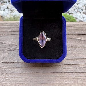 May include: A silver ring featuring a marquise-cut purple gemstone set in a decorative metal band. The ring is displayed inside a square, royal blue velvet-lined jewelry box with a hinged lid, showcasing the stone's faceted surface and metallic detail.