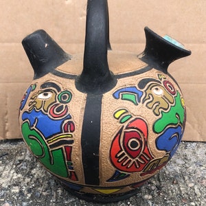 May include: A brown ceramic pitcher with a black handle and spout. The pitcher is decorated with colorful geometric patterns and stylized figures.