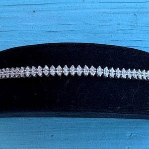 May include: A silver-toned tennis bracelet featuring a continuous row of small, clear, round-cut stones set in a metallic band. The bracelet is displayed horizontally, showcasing a uniform arrangement of sparkling stones along its entire length.