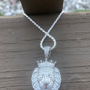 May include: A silver chain necklace with a pendant featuring a lion's head with a crown, all covered in sparkling white cubic zirconia stones.