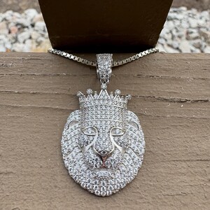 May include: Silver-toned pendant necklace featuring a lion's head wearing a crown, encrusted with small, sparkling stones. The pendant hangs from a delicate chain. The necklace is displayed on a wooden surface.