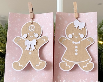 May include: Two pink gift bags with gingerbread man and woman cutouts, held by wooden clips. The gingerbread figures have white accents and bows. The bags have a snowflake pattern and the text "MyPaperPlanet".