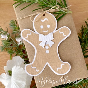 May include: A brown gingerbread man gift tag with white icing details, including a bow tie and buttons. The tag is tied with twine and is on a brown paper gift bag.