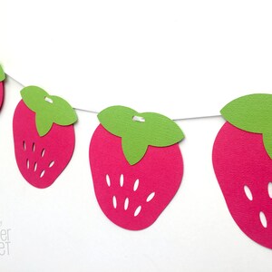May include: A string of paper strawberry cutouts, each with a green leaf, strung together with white thread. The strawberries are pink and have white seeds.