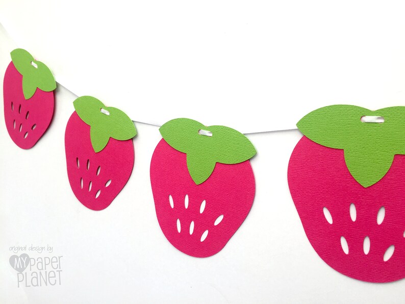 May include: A string of paper strawberry cutouts, each with a green leaf, strung together with white thread. The strawberries are pink and have white seeds.