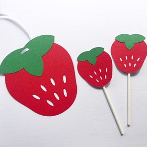 May include: Three paper cutouts of strawberries in different sizes. The largest strawberry is red with white seeds and a green leafy top. The two smaller strawberries are on sticks and are also red with white seeds and green leafy tops.