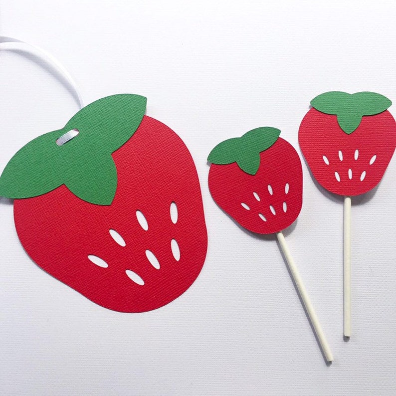 May include: Three paper cutouts of strawberries in different sizes. The largest strawberry is red with white seeds and a green leafy top. The two smaller strawberries are on sticks and are also red with white seeds and green leafy tops.