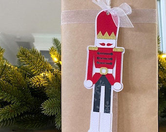 May include: A gift wrapped in brown paper with a sheer ribbon, adorned with a red and gold nutcracker tag. The tag features a soldier design with a red hat, gold accents, and white boots, perfect for holiday gifting.
