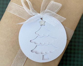 May include: A white, circular gift tag with a cut-out Christmas tree design, adorned with a small, sparkly star. The tag is tied with a sheer, light-colored ribbon and is attached to a wrapped gift. The gift is wrapped in brown paper.