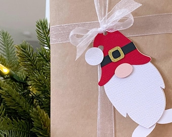 May include: A brown paper gift bag with a white ribbon and a gnome-shaped gift tag. The gnome is wearing a red hat with a black belt and a white beard.