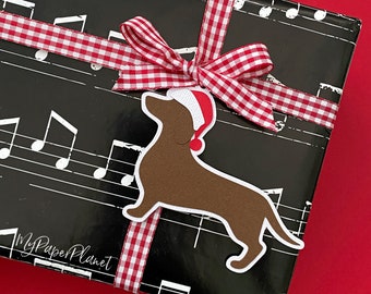 May include: A wrapped gift with black paper printed with white musical notes. A red and white checkered ribbon is tied around the gift, with a bow on top. A brown dachshund wearing a Santa hat is attached to the ribbon.