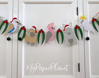 May include: A Christmas banner featuring Australian animals wearing Santa hats. The banner includes a kookaburra, a platypus, a pink cockatoo, a koala, and a cockatoo. The banner is decorated with green eucalyptus leaves and red berries.