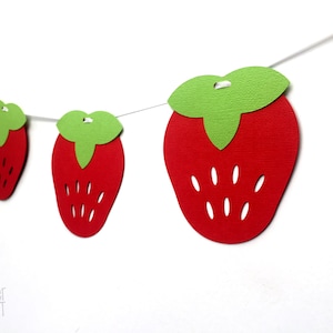 May include: A red and green paper strawberry garland with white string. The garland is made of paper cutouts of strawberries with green leaves. The strawberries have white seeds.