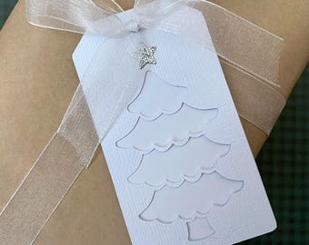 May include: A white gift tag with a cut-out Christmas tree design and a silver star embellishment. The tag is tied with a sheer white ribbon and is wrapped around a brown package. The tag has the text "MyPaperPlanet" at the bottom.