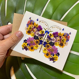 May include: A white greeting card featuring a butterfly design made of pressed flowers in shades of yellow, purple, and blue. The card is held with a brown envelope. The background includes a large green leaf.