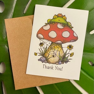 May include: A whimsical greeting card featuring a cartoon frog wearing a cowboy hat, resting on a red and white mushroom. The card includes the text "Thank You!" and is accompanied by a brown envelope. The illustration has a spider web and flowers.