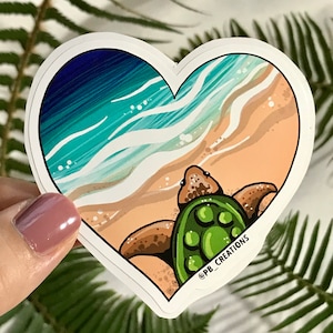 May include: Heart-shaped sticker featuring a sea turtle on a beach. The design includes a green turtle, tan sand, and blue and turquoise ocean waves. The sticker has a white border and the text "@PB_CREATIONS".