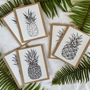 May include: A collection of blank greeting cards featuring detailed black and white pineapple illustrations. The cards have a white interior and a brown border. Fern leaves are in the background.