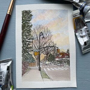 May include: Watercolor painting of a street scene with a crosswalk, streetlights, and trees. The artwork features a muted color palette with shades of blue, yellow, and brown. Paint tubes and a paintbrush are in the foreground.