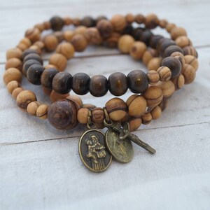 May include: Three olive wood beaded bracelets with a brown wooden bead bracelet in the center. The bracelets have a small gold-toned charm with a religious image and a small gold-toned cross charm.