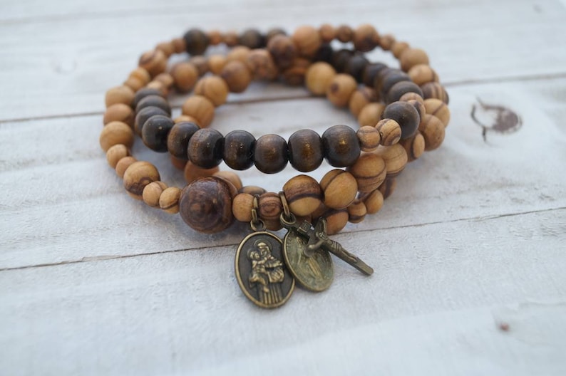 May include: Three olive wood beaded bracelets with a brown wooden bead bracelet in the center. The bracelets have a small gold-toned charm with a religious image and a small gold-toned cross charm.