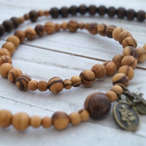 May include: A wooden beaded necklace with a brown and natural wood color scheme. The necklace has a small metal charm with a religious image.