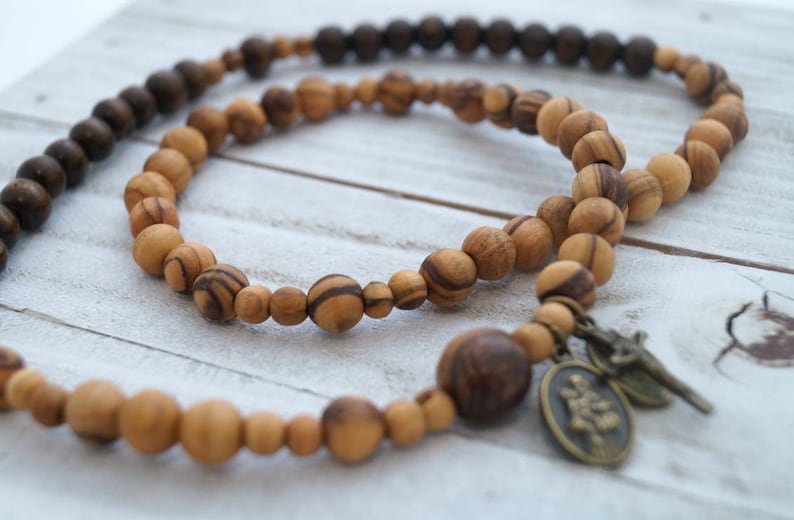 May include: A wooden beaded necklace with a brown and natural wood color scheme. The necklace has a small metal charm with a religious image.