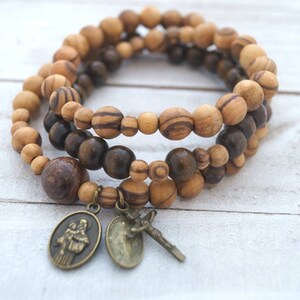 May include: A triple-strand bracelet made of brown and light brown wooden beads with two metal charms. One charm features a religious figure and the other charm features a cross.