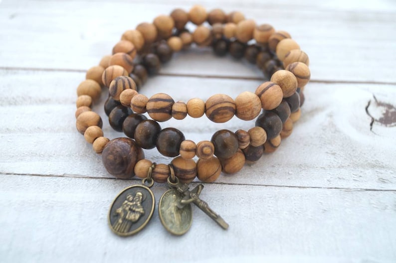May include: A triple-strand bracelet made of brown and light brown wooden beads with two metal charms. One charm features a religious figure and the other charm features a cross.