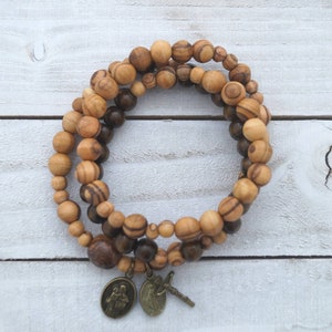 May include: A double-strand olive wood bracelet with brown and light brown beads. The bracelet has two metal charms, one with a religious image and the other with a cross.