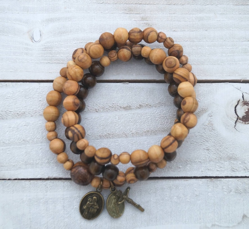 May include: A double-strand olive wood bracelet with brown and light brown beads. The bracelet has two metal charms, one with a religious image and the other with a cross.