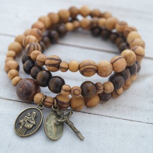 May include: A triple-strand bracelet made of brown and light brown wooden beads with two metal charms. One charm features a depiction of two figures, and the other charm features a figure with a cross.