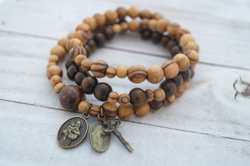 May include: A triple-strand bracelet made of brown and light brown wooden beads with two metal charms. One charm features a depiction of two figures, and the other charm features a figure with a cross.