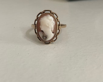 May include: A gold ring with a cameo featuring a woman's profile. The cameo is set in a gold oval frame with a delicate design. The woman is wearing a necklace with a small diamond pendant.