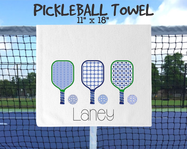 Op de afbeelding: Witte pickleball handdoek met drie blauwe en witte gepatroneerde pickleball rackets en drie blauwe en witte gepatroneerde pickleballen. De handdoek heeft de naam "Laney" erop gedrukt. De handdoek is 28 cm bij 46 cm.
