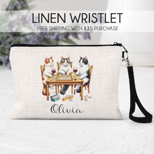 May include: Linen wristlet with a black strap. The wristlet features a design of three cats playing dominoes with glasses of wine. The text "Olivia" is printed on the wristlet.