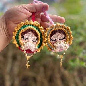 May include: Two handmade crochet ornaments featuring a face with closed eyes and clasped hands. The ornaments have a gold, green, and yellow halo design. Each ornament has a gold rosary with a cross. One has a red loop, the other a pink loop.