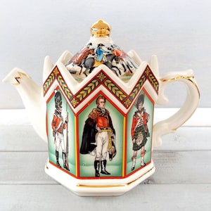 May include: A white ceramic teapot with a crown-shaped lid and gold accents. The teapot features illustrations of soldiers in red coats and a scene of riders on horseback. The handle and spout are also white with gold trim.