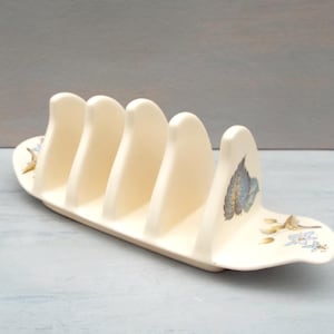 Peut inclure: Un porte-toast en céramique blanche avec quatre fentes et un motif floral. Le porte-toast est en forme de bateau et a une poignée incurvée.