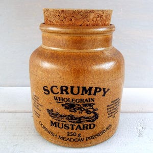 May include: A brown ceramic jar with a cork stopper, labelled "SCRUMPY WHOLEGRAIN MUSTARD." The label features a black illustration of a landscape and text. The jar contains 250g of mustard and is from Cornish Meadow Preserves.
