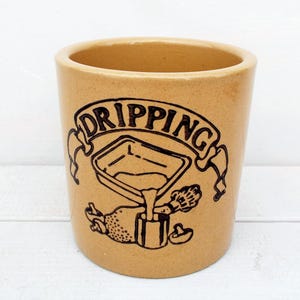 May include: Tan ceramic crock with a black graphic of a pouring vessel and the word "DRIPPING" in a banner. The crock has a cylindrical shape and a wide opening, suitable for kitchen use.