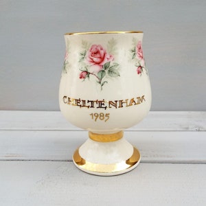 May include: A white ceramic goblet with a gold rim and base. The goblet is decorated with pink roses and green leaves. It has the words "Cheltenham 1985" written in gold on the front.